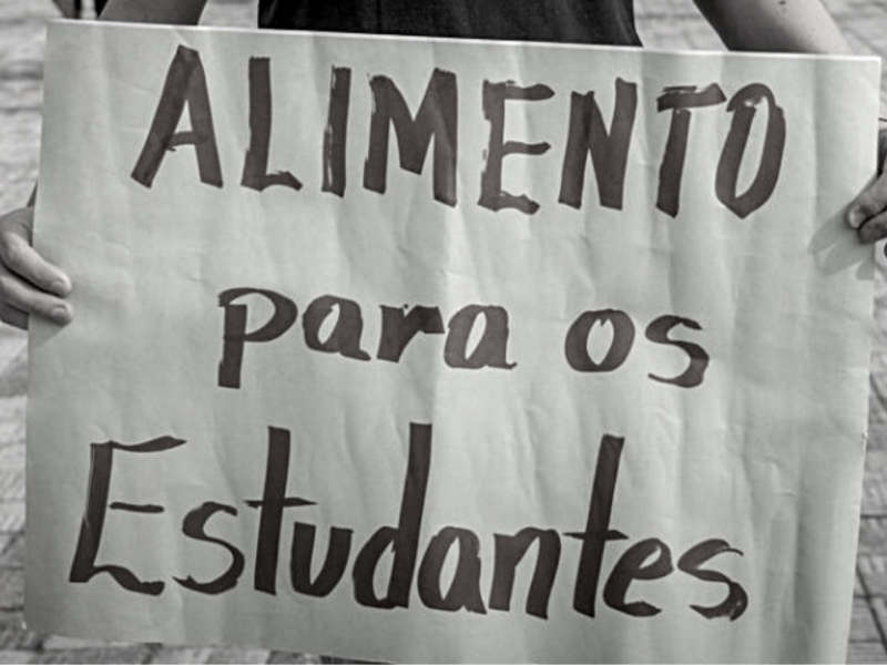 Foto tem pessoa segurando cartaz onde se vê texto "Alimento para os estudantes"