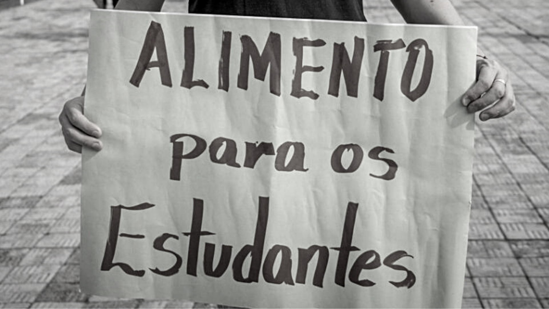 Foto tem pessoa segurando cartaz onde se vê texto "Alimento para os estudantes"